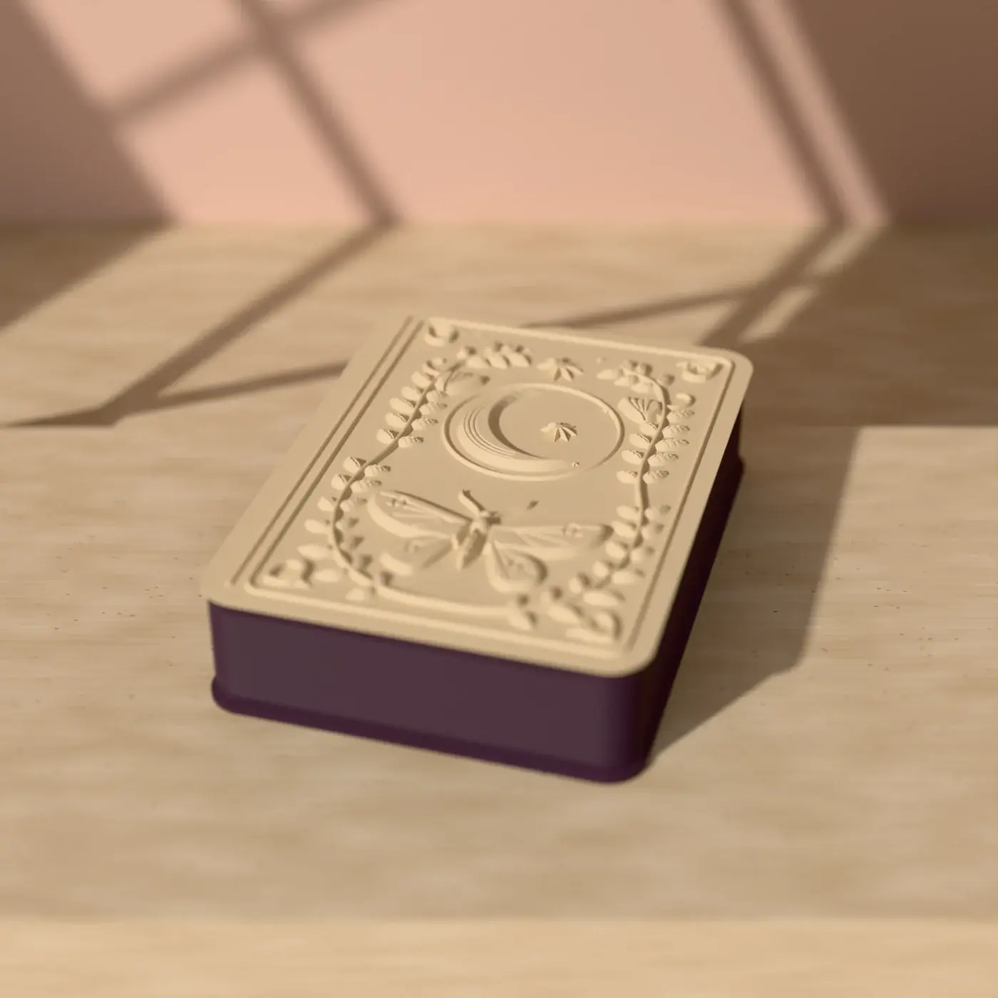 The Midnight Grimoire box on a sunlit table: a plum book-shaped box under an ivory cover sculpted with a crescent moon, luna moth and leafy wreath.