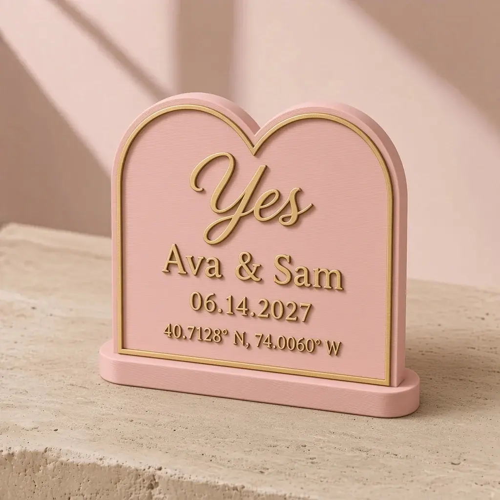 Blush heart-arched plaque with gold lettering reading Yes, Ava & Sam, 06.14.2027 and map coordinates, on a small stand.