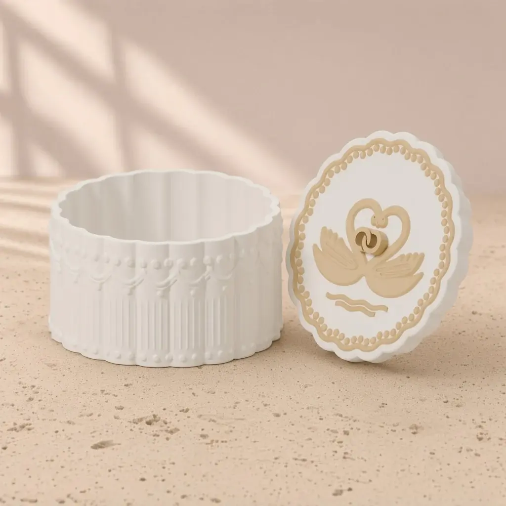 Design image of the swan heart box: a scalloped ivory lace box beside its lid with two gold swans forming a heart around interlocked rings.